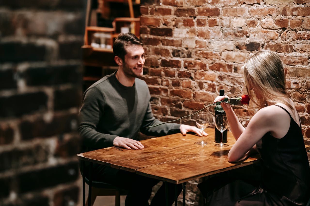 Young couple in elegant clothes sitting in light cafeteria at wooden table near brick wall and having romantic dinner with red rose and wine near glasses