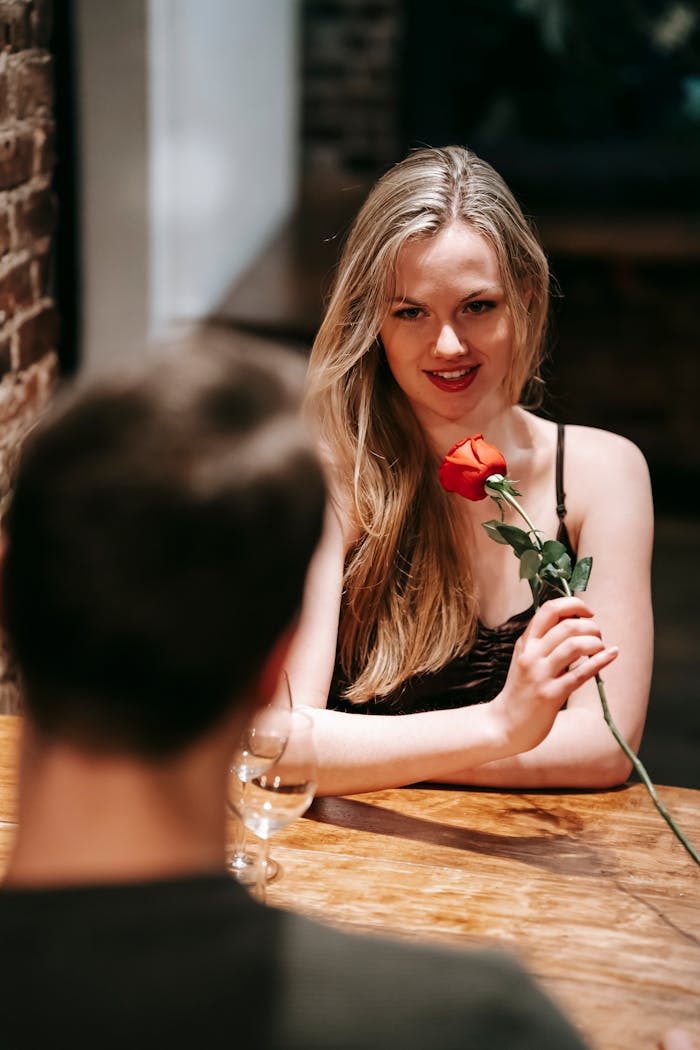 Young couple in elegant clothes sitting at table in light cafe while enjoying romantic dinner with wineglasses and red rose
